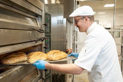 Kuvassa hymyilevä Samuli Honkanen leipomo Juuresta ottaa paistouunista valmiita leipiä ulos.